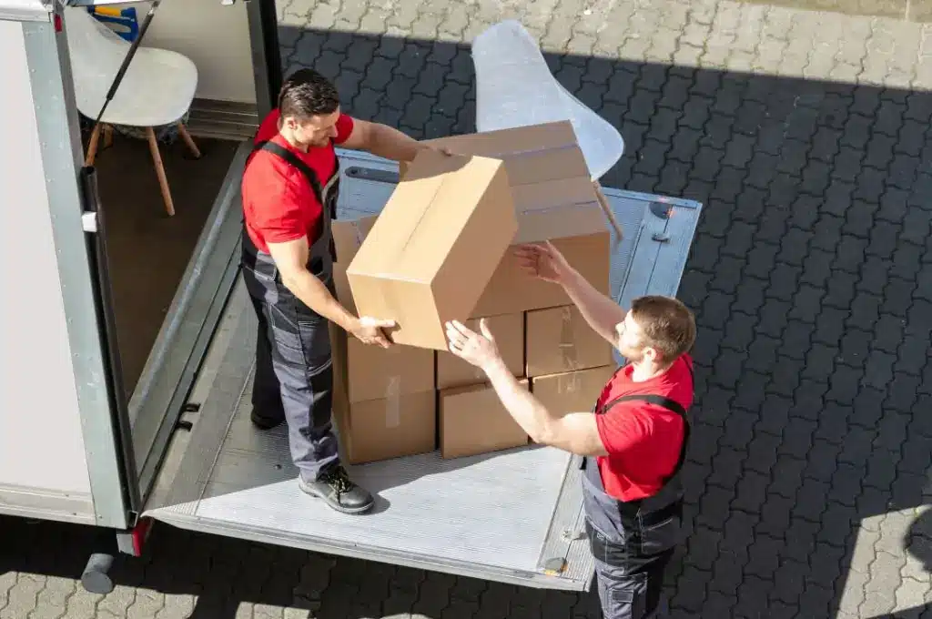 Deux déménageurs en salopette rouge et grise chargent des cartons marrons dans un camion de déménagement, travaillant ensemble.