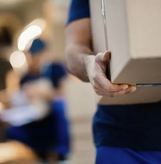 Livreur en uniforme bleu portant un carton. Main tenant le colis avec d'autres travailleurs en arrière-plan flou. Scène de logistique ou déménagement.