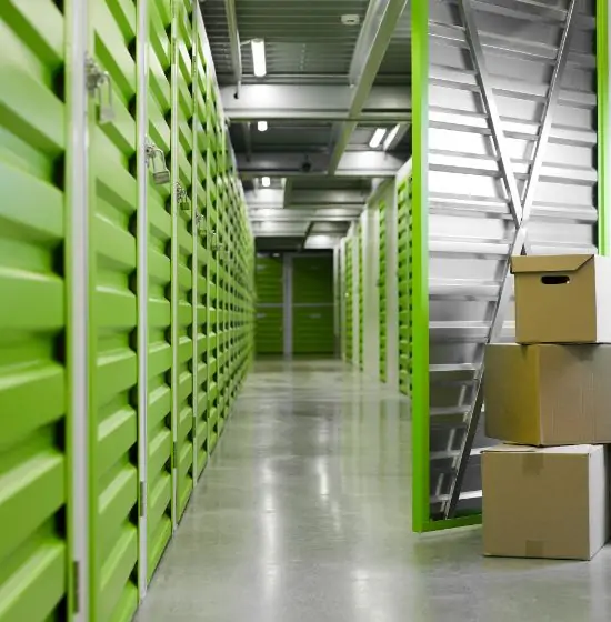 Allée d'un centre de self-stockage moderne avec des unités vertes lumineuses verrouillées et des cartons empilés sur la droite.